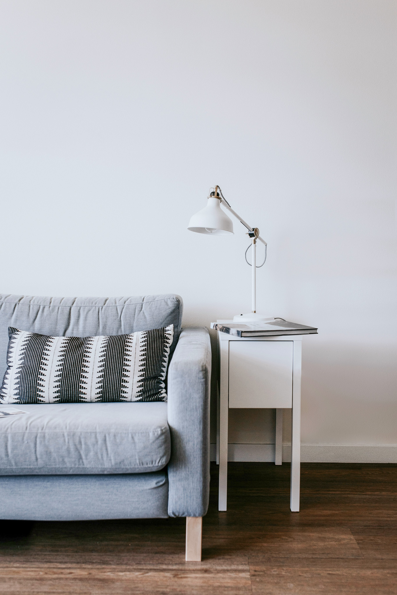 Ein graues Sofa mit gemusterten Kissen steht neben einem kleinen weißen Beistelltisch, auf dem eine weiße Schreibtischlampe und ein geschlossener Laptop stehen, vor einer schlichten weißen Wand auf einem Holzboden.