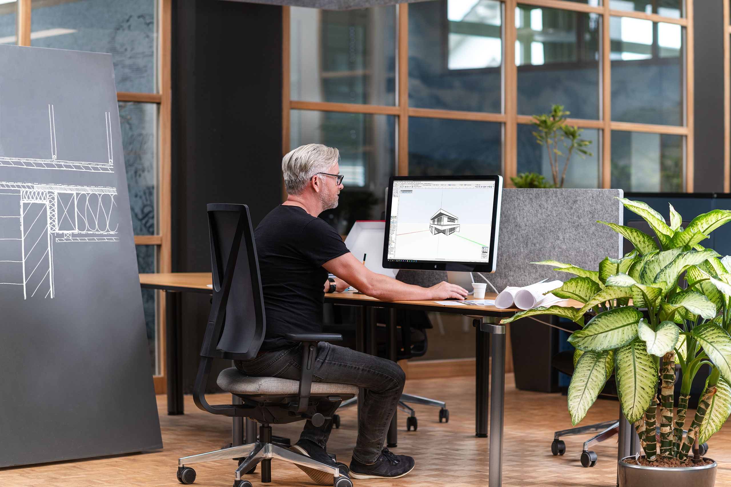 Ein Mann sitzt an einem Schreibtisch in einem Büro und arbeitet am Computer an Architekturentwürfen. In der Nähe stehen eine große Anlage und ein Zeichenbrett.