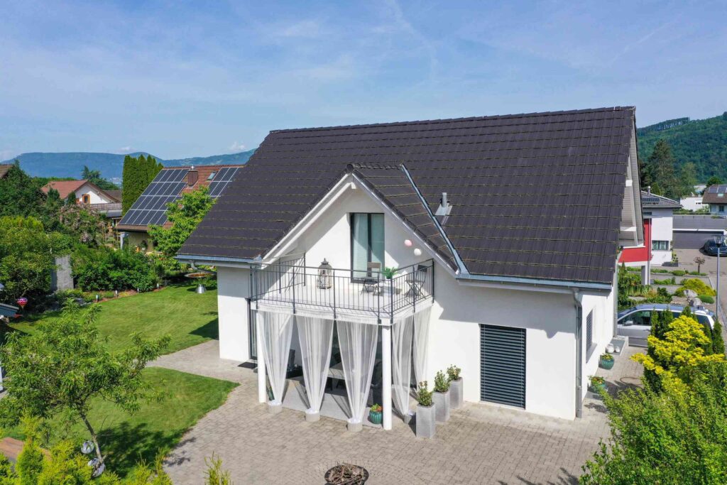 Luftaufnahme eines weißen Hauses mit dunklem Dach, einem kleinen Balkon, einer Terrasse und umgebendem Grün. Im Hintergrund sind Sonnenkollektoren auf einem Nachbarhaus zu sehen.