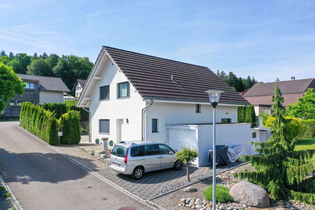 Ein weißes zweistöckiges Haus mit dunklem Dach und angebauter Garage. In der Einfahrt parkt ein silberner Minivan. Die Straße ist von Grün gesäumt und die Nachbarhäuser sind sichtbar.