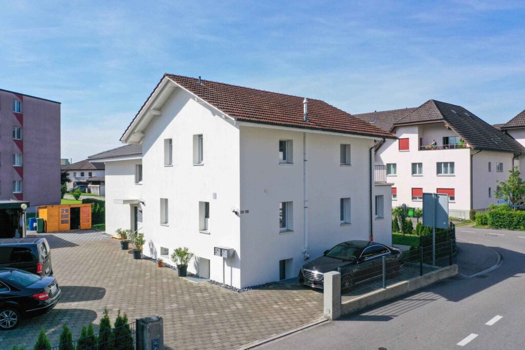 An einer Straßenecke in einem Wohngebiet steht ein weißes, zweistöckiges Haus mit rotem Ziegeldach, umgeben von Autos und einer kleinen gepflasterten Auffahrt.