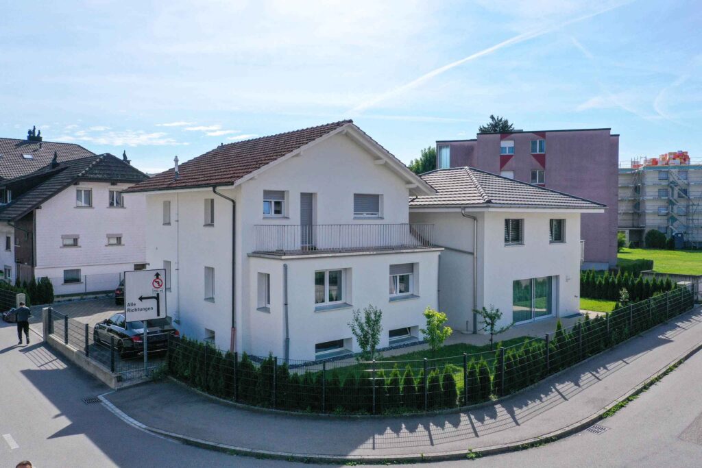 Ein weißes zweistöckiges Haus mit eingezäuntem Garten auf einem Eckgrundstück, umgeben von ähnlichen Wohngebäuden und darüber klarem blauen Himmel.