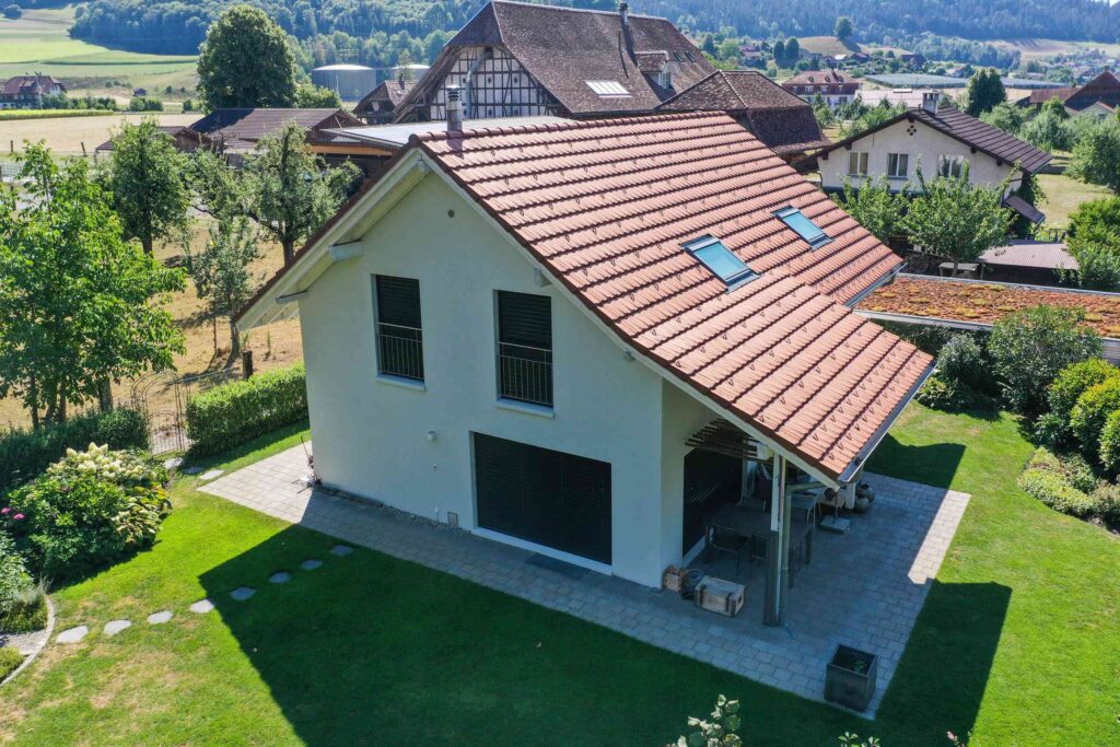 Ein kleines Haus mit einem roten Ziegeldach und zwei Oberlichtern, umgeben von einem Garten und angrenzend an Felder und andere Häuser.