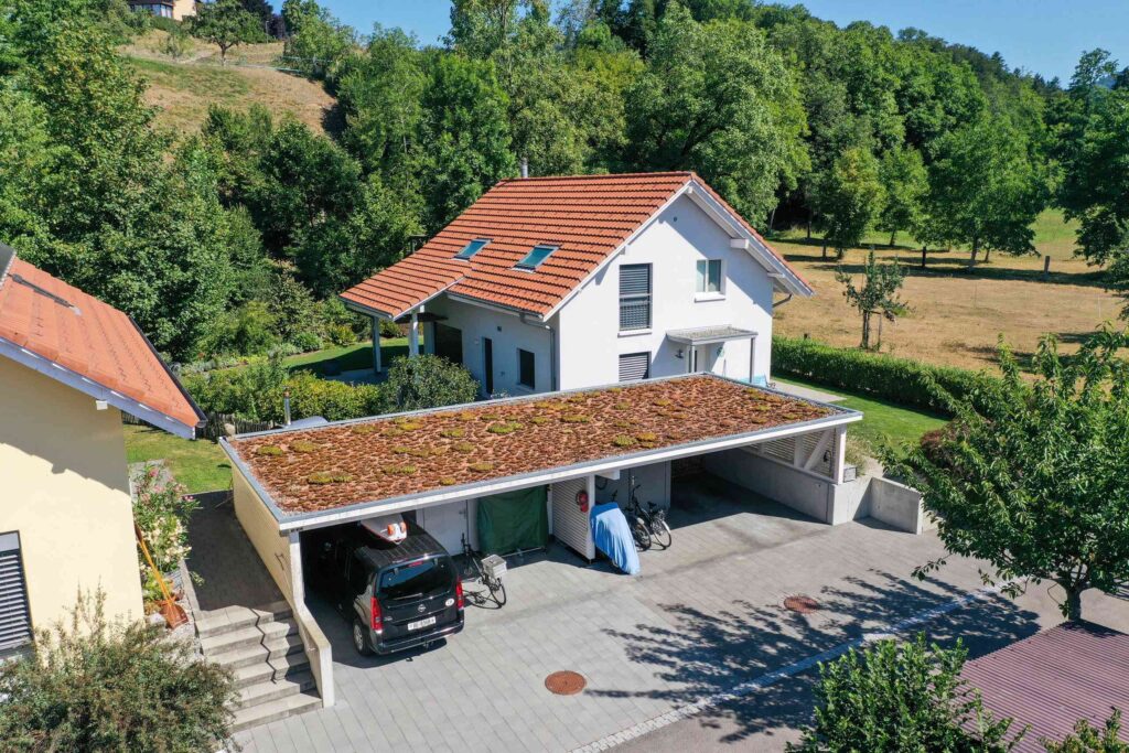 Ein weißes Haus mit rotem Dach ist von viel Grün umgeben. Davor befindet sich ein Carport mit Gründach, unter dem Fahrzeuge und Fahrräder abgestellt werden.