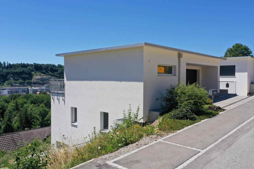 Modernes weißes einstöckiges Haus mit Flachdach, an einem grünen Hang gelegen, gegenüber einer asphaltierten Straße.