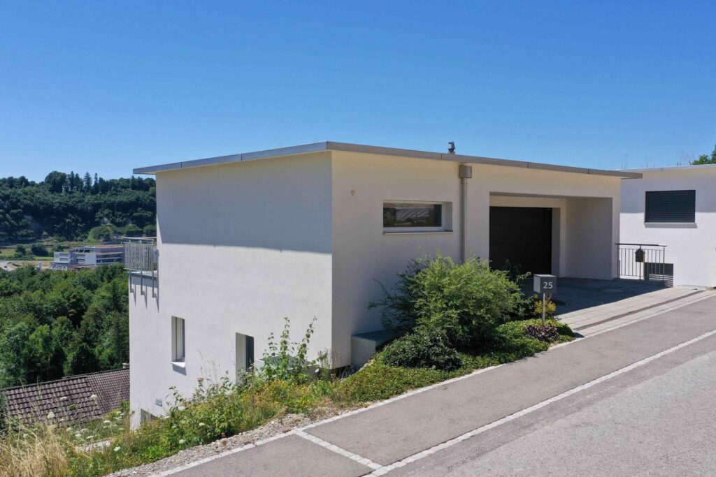Modernes weißes Haus mit Flachdach an einem Hang gelegen, mit Auffahrt und Grünfläche. Straße im Vordergrund, Hügel und Bäume im Hintergrund unter einem klaren blauen Himmel.