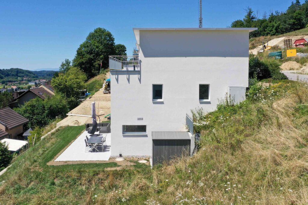 Ein modernes weißes Haus mit Flachdach, gelegen auf einem abschüssigen Grasgelände, mit einer Terrasse mit Gartenmöbeln und umgeben von Bäumen und Nachbarhäusern.