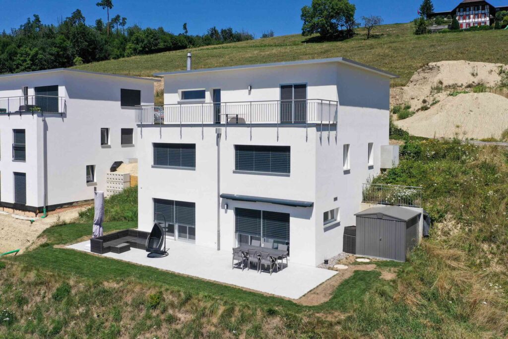 Modernes weißes Haus am Hang mit zwei Etagen, Balkonen und einem Außensitzbereich auf einer gepflasterten Terrasse. Angrenzend an ein ähnliches Haus und umgeben von Grasgelände.