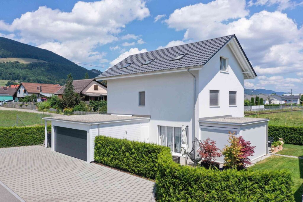 Zweistöckiges weißes Haus mit grauem Dach, umgeben von einer Hecke, einer Auffahrt und einer Garage, in einer Vorstadtgegend mit Bergen im Hintergrund unter einem teilweise bewölkten Himmel.