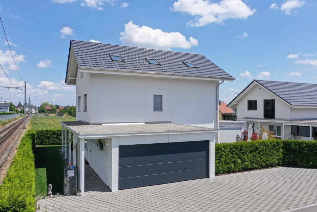 Ein modernes zweistöckiges weißes Haus mit einer dunkelgrauen Garage neben einer Bahnstrecke. Die Auffahrt ist mit hellgrauen Fliesen gepflastert und Sträucher säumen die Seite des Hauses.
