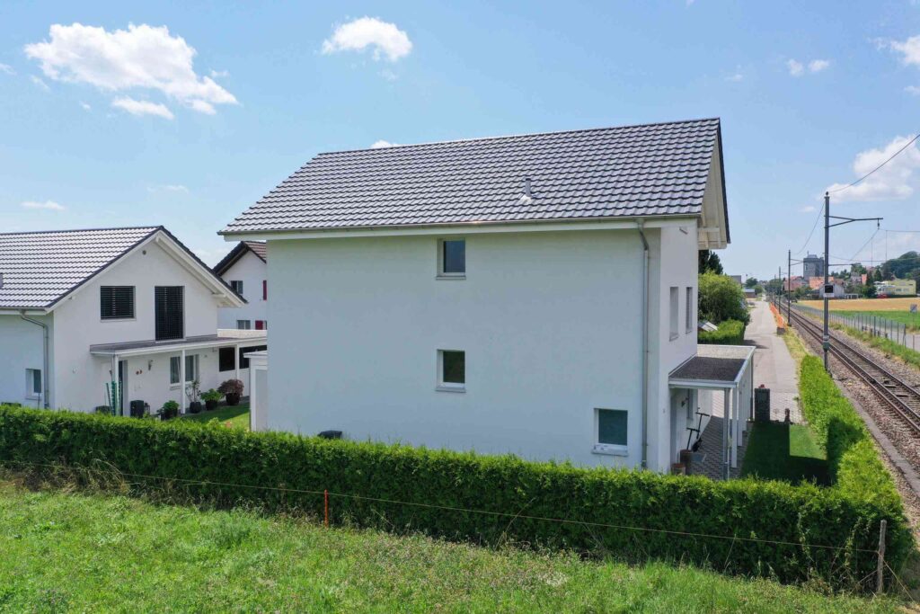 Modernes weißes zweistöckiges Haus mit Ziegeldach, neben einer Bahnstrecke und umgeben von Grün, unter einem teilweise bewölkten Himmel.