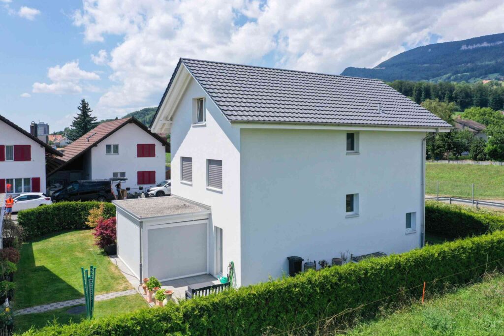 Ein weißes zweistöckiges Haus mit grauem Dach liegt in einer grünen Vorstadtgegend. Es verfügt über einen kleinen Garten, Hecken und Nachbarhäuser. Im Hintergrund sind Hügel und ein teilweise bewölkter Himmel zu sehen.