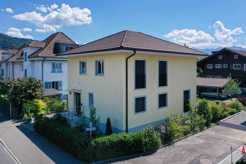 Ein zweistöckiges gelbes Haus mit einem braunen Ziegeldach steht auf einem Eckgrundstück, umgeben von Grün, unter einem klaren blauen Himmel.