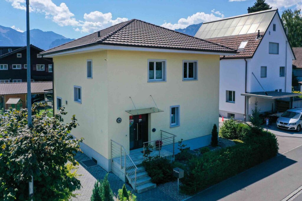 Zweistöckiges gelbes Haus mit Ziegeldach, Vordertreppe und kleinen Sträuchern. Auf dem Haus dahinter ist ein Solarpanel sichtbar.