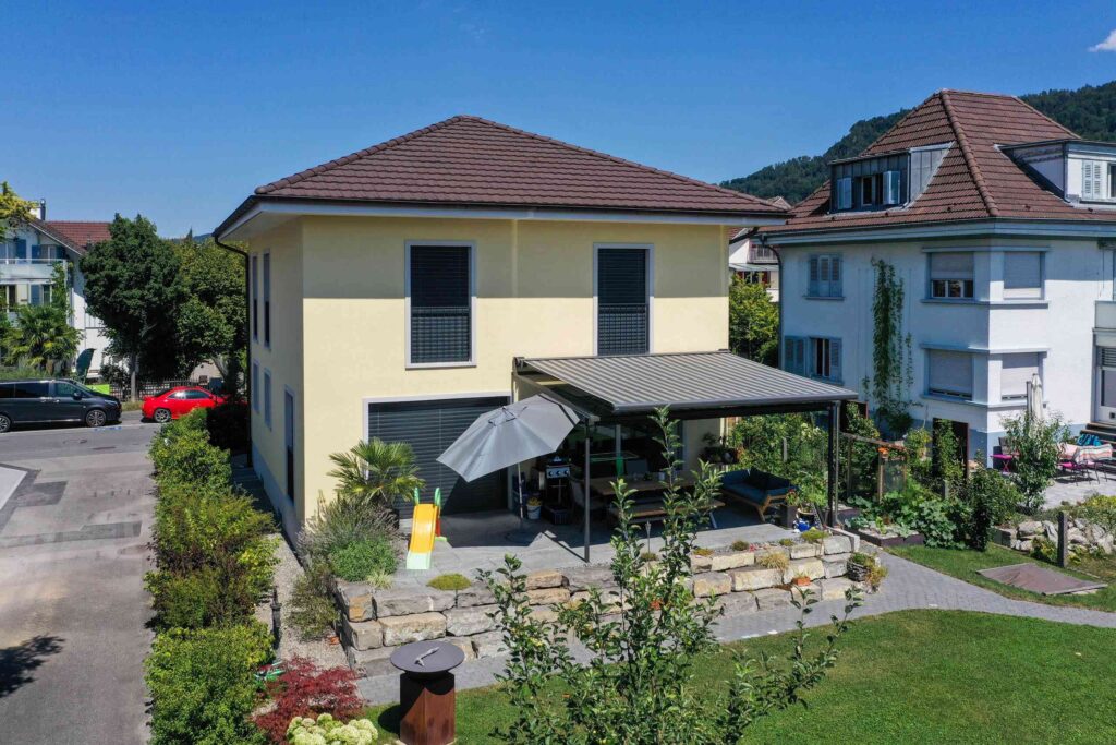 Ein zweistöckiges gelbes Haus mit einem braunen Dach, einer überdachten Terrasse und einem Garten. In der Nähe stehen andere Häuser und ein geparktes rotes Auto. Der Himmel ist klar und sonnig.