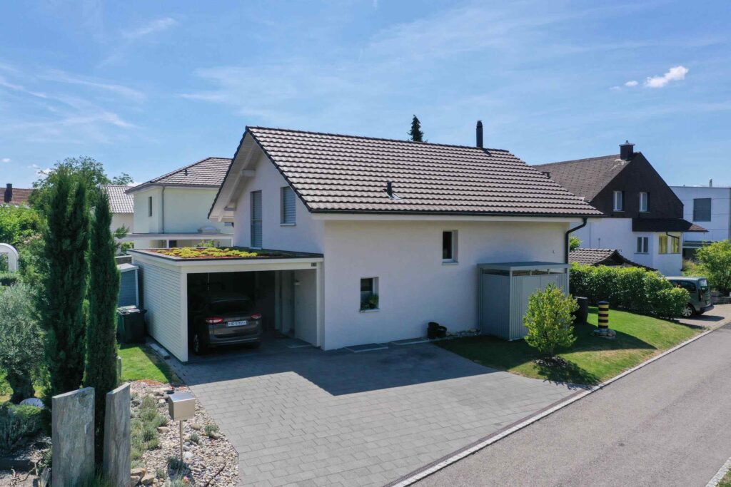 Ein modernes, zweistöckiges weißes Haus mit Ziegeldach, angebauter Garage und kleinem Garten, gelegen in einer ruhigen Straße unter einem klaren blauen Himmel.