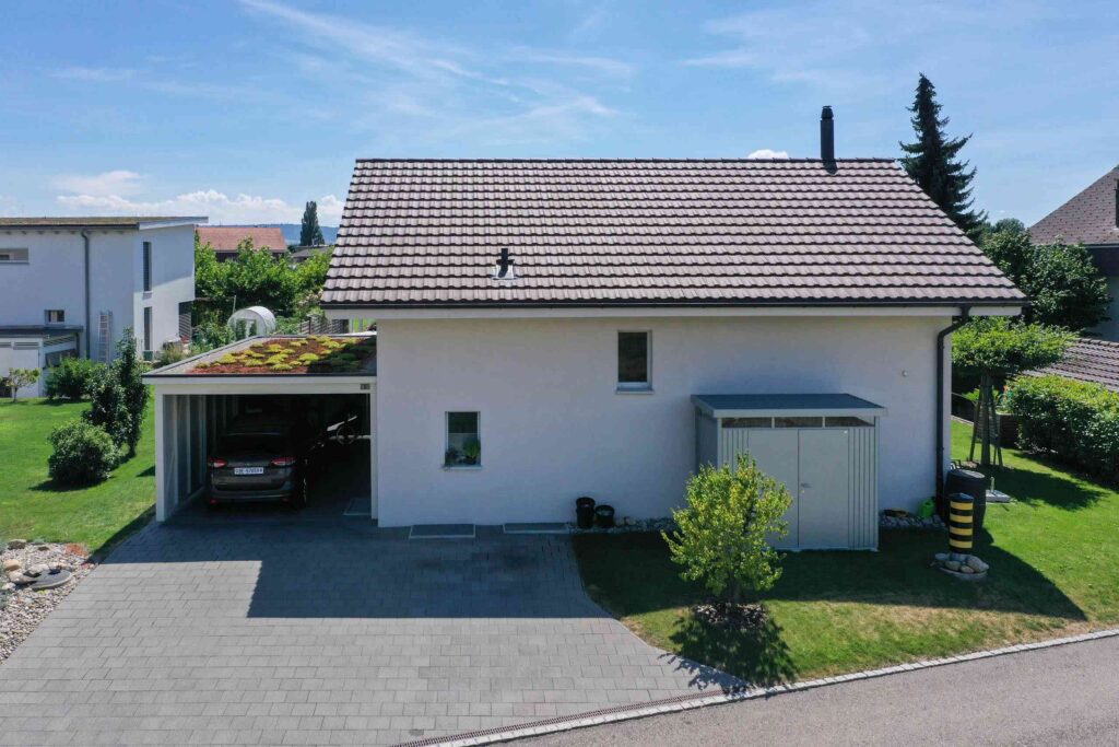 Ein modernes Haus mit Satteldach und angeschlossenem Carport auf der linken Seite, gelegen in einem gepflegten Hof mit Grünflächen, unter einem klaren blauen Himmel.