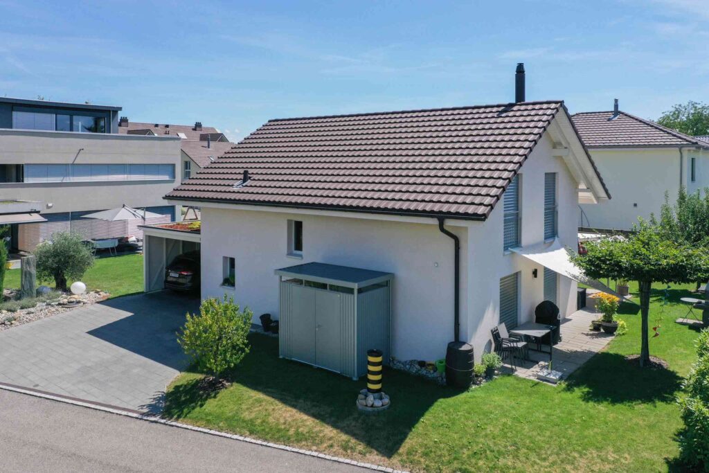 An einem sonnigen Tag steht ein weißes zweistöckiges Haus mit braunem Ziegeldach, Garage und kleinem Gartenbereich in einem Wohnviertel.