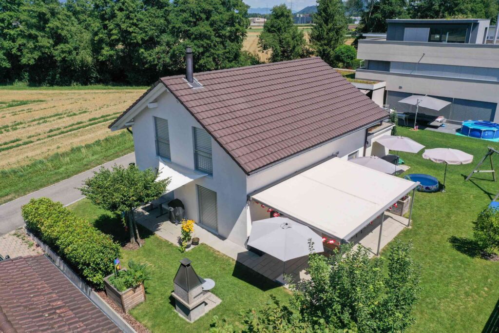 Luftaufnahme eines weißen Hauses mit braunem Dach, umgeben von Grün. Der Hinterhof verfügt über eine gepflasterte Terrasse mit Sonnenschirmen und einem kleinen runden Pool. In der Nähe befindet sich ein zweites modernes Gebäude.