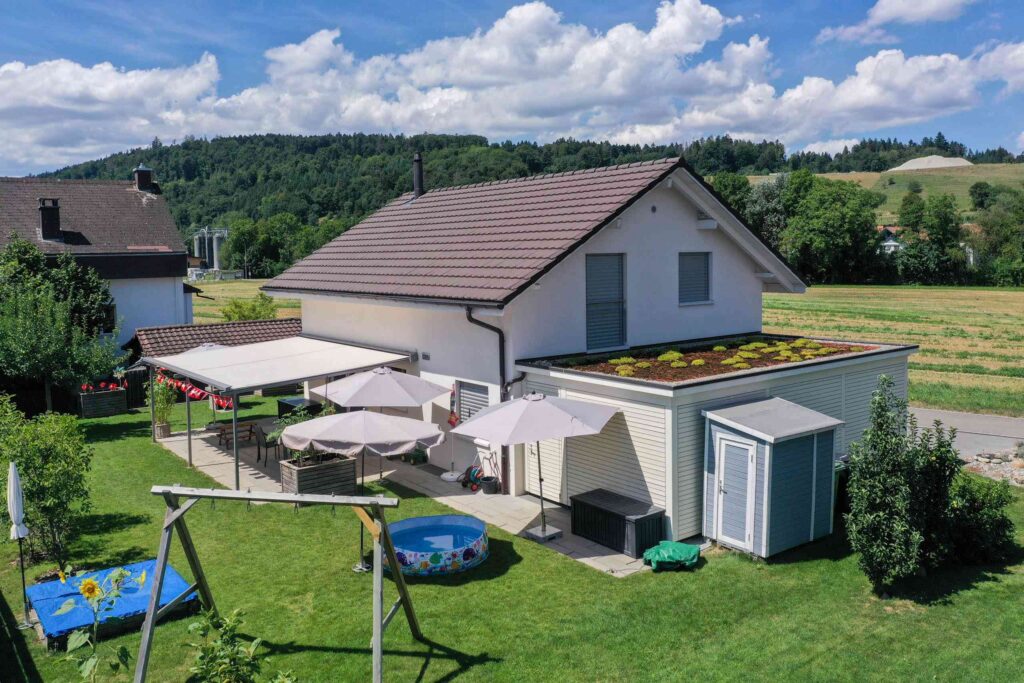 Ein modernes Haus mit rotbraunem Dach, Sonnenkollektoren und einem grünen Dachabschnitt. Der Hinterhof verfügt über eine Pergola, ein aufblasbares Schwimmbecken, Sonnenschirme und eine Schaukel vor einer ländlichen Landschaft.