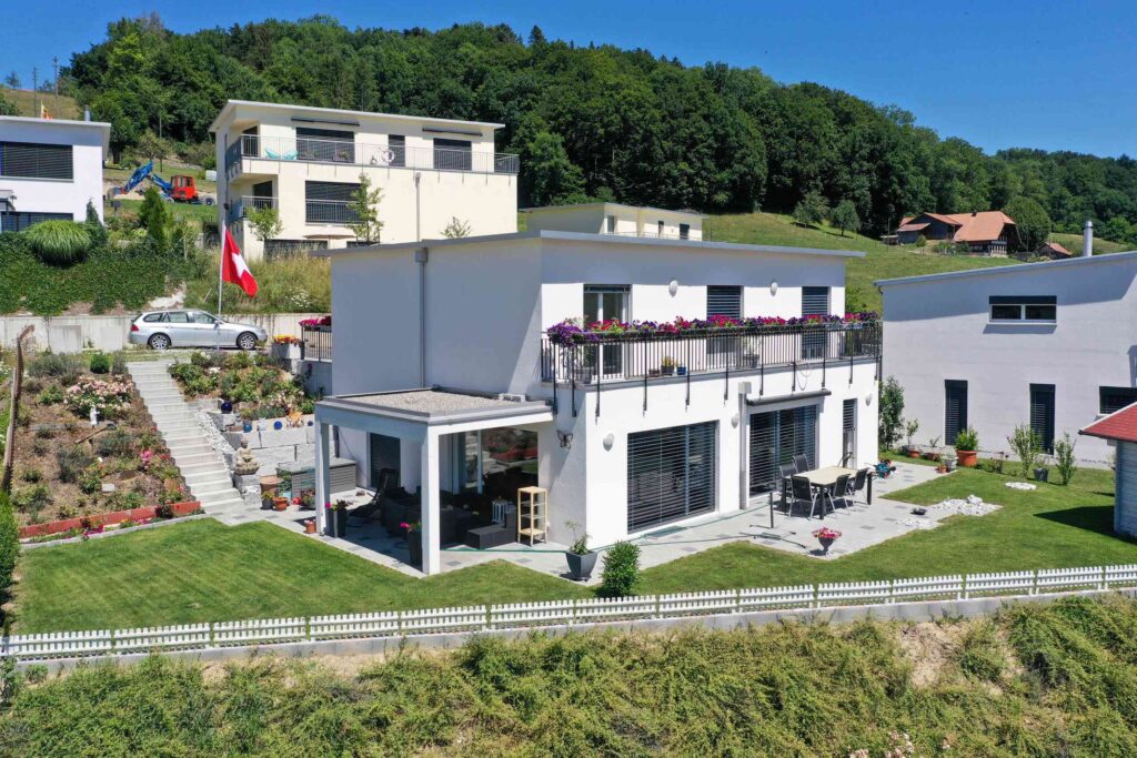 Modernes weißes Haus mit Balkon, gepflegtem Rasen und Schweizer Flagge im Garten, umgeben von anderen Häusern und Bäumen in einer hügeligen Gegend.