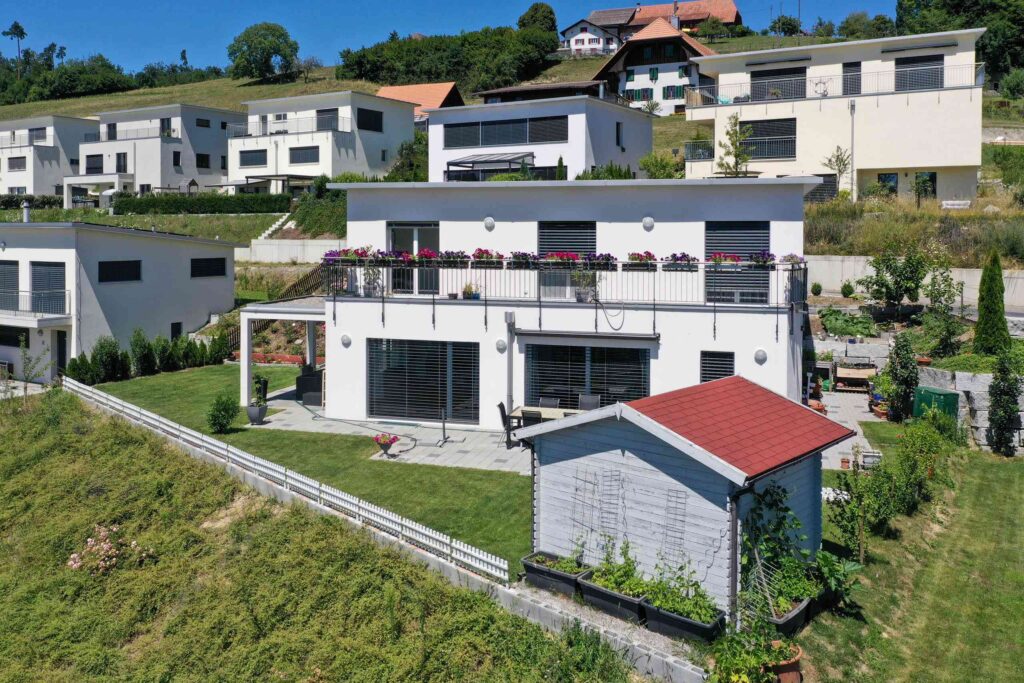 Ein zweistöckiges weißes Haus mit einer großen Terrasse und Topfblumen, gelegen auf einem grünen Rasen, mit benachbarten modernen Häusern im Hintergrund.