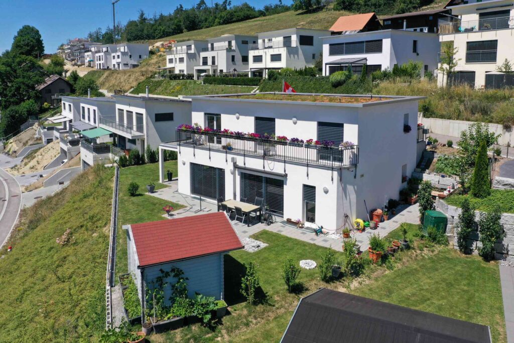 Ein modernes weißes Haus mit Flachdach und einem großen, mit Blumen geschmückten Balkon. Im Hinterhof steht ein Schuppen mit rotem Dach, umgeben von einem gepflegten Garten. Auf einem Hügel sind weitere Häuser zu sehen.
