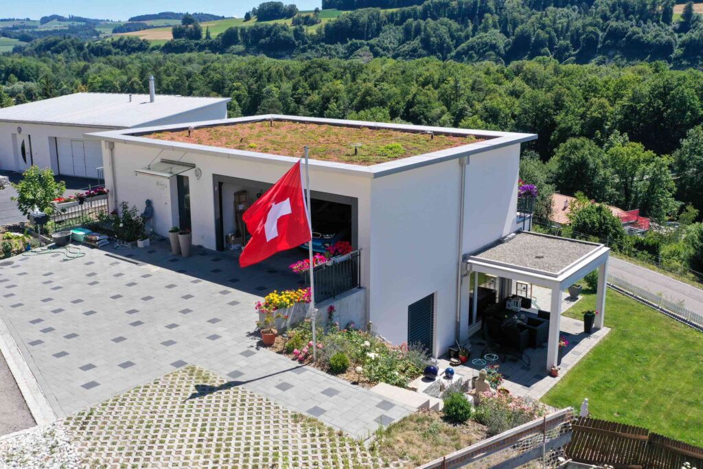 Modernes weißes Haus mit Flachdach und Solarmodulen, umgeben von Grün. Vorne prangt prominent eine Schweizer Flagge. Gepflasterte Terrasse mit Blumen und einem kleinen überdachten Bereich an der Seite.