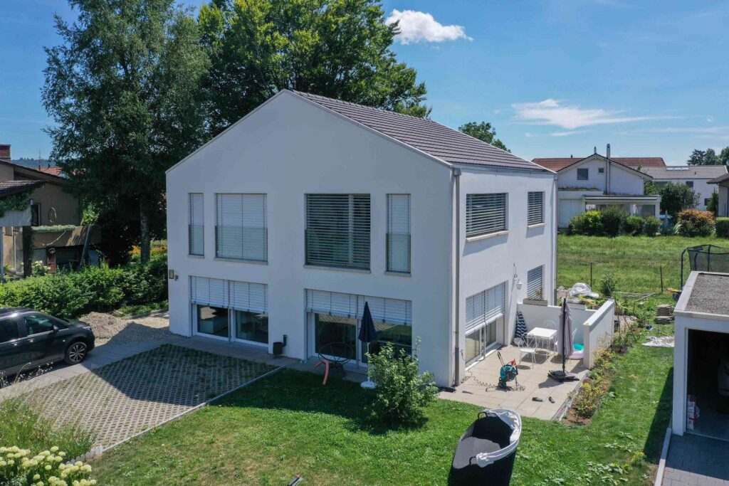 Ein modernes zweistöckiges weißes Haus mit großen Fenstern, einer Terrasse und einem kleinen Hinterhof. Auf dem Grundstück steht ein geparktes Auto und es gibt viel Grün.