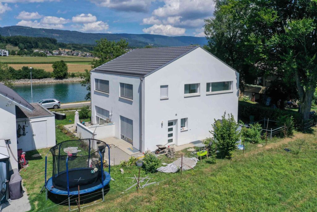 Ein modernes weißes zweistöckiges Haus mit einem Trampolin im Hinterhof, in der Nähe eines Flusses gelegen und von viel Grün umgeben.