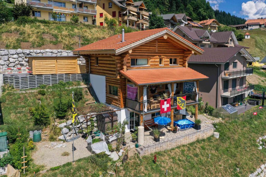 Ein Holzhaus am Hang mit einer Schweizer Flagge und anderen Bannern. Es hat eine kleine Terrasse mit Gartenmöbeln und ist von anderen Häusern umgeben.