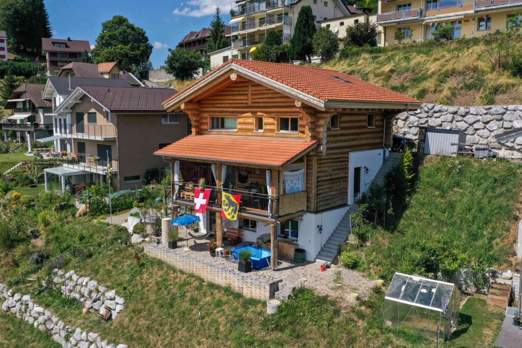 Ein Holzhaus mit einer Terrasse, auf der Schweizer und kantonale Flaggen zu sehen sind, liegt an einem Hang, umgeben von Grün und umliegenden Gebäuden.