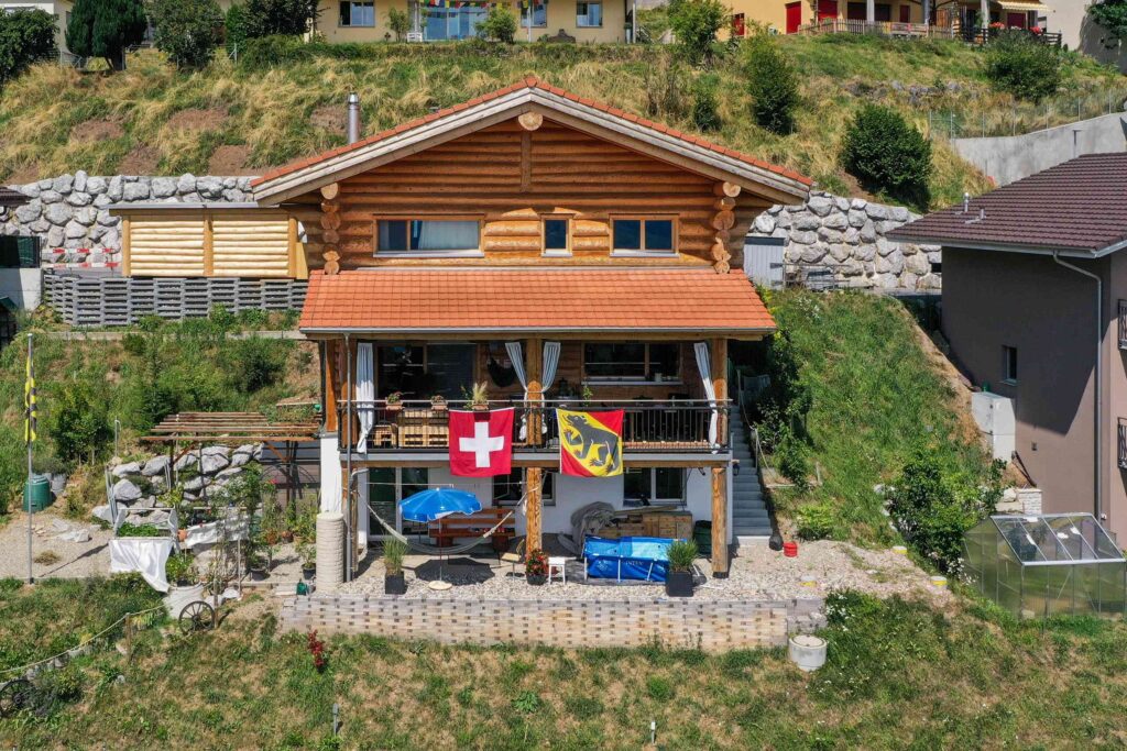 Ein Holzhaus mit einer Terrasse, auf der die Schweizer und Berner Flaggen zu sehen sind. Stufen führen zu einem Garten und auf einem grasbewachsenen Hügel sind benachbarte Häuser zu sehen.