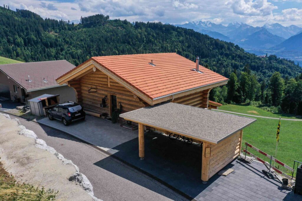 Eine Blockhütte mit rotem Dach und Carport liegt eingebettet in einer Berglandschaft. In der Nähe parkt ein Auto und im Hintergrund bietet sich eine malerische Aussicht auf Hügel und Berge.