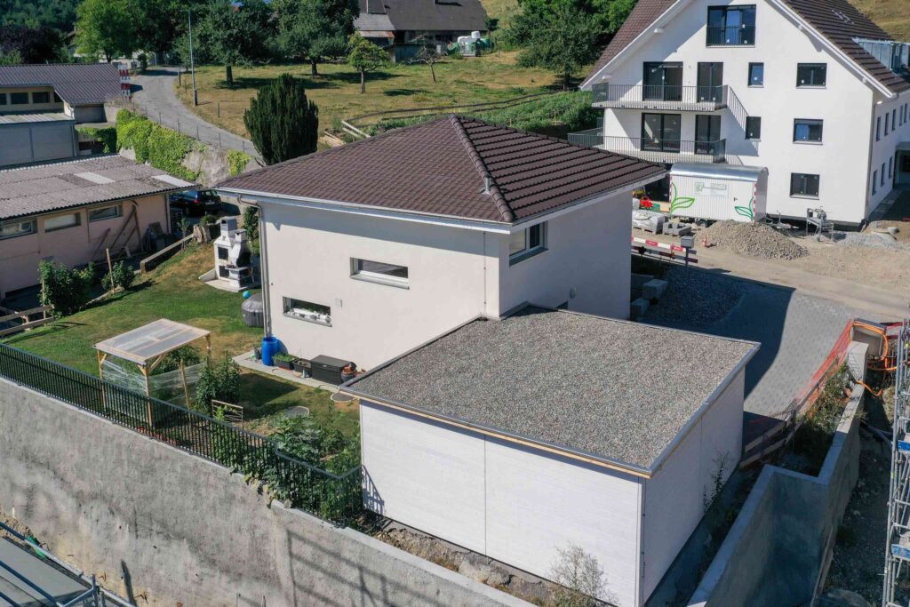 Luftaufnahme eines Wohngebiets mit einem weißen Haus, einer Garage, einem kleinen Schuppen und einem mehrstöckigen Gebäude. Grüne Rasenflächen und Einfahrten umgeben die Gebäude.