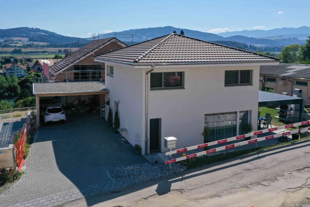 Zweistöckiges weißes Haus mit Ziegeldach, Carport für ein Fahrzeug und angelegtem Vorgarten. Im Vordergrund ist eine rot-weiße Bauabsperrung zu sehen. Im Hintergrund Berge.