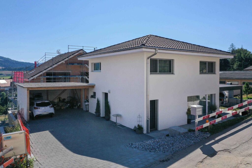 Ein modernes zweistöckiges weißes Haus mit Ziegeldach, einem Carport mit geparktem Fahrzeug und einem im Bau befindlichen Nachbarhaus mit Gerüsten und Leitern.