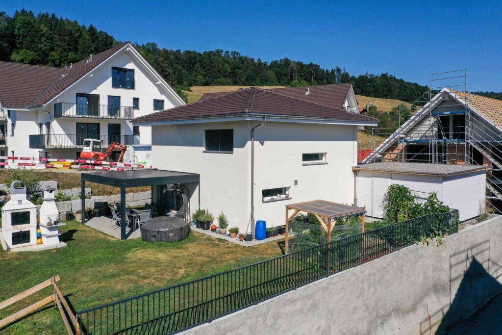 In einem Vorstadtgarten sind ein weißes zweistöckiges Haus, eine überdachte Terrasse, ein Whirlpool und ein eingezäunter Gartenbereich zu sehen. Im Hintergrund sind weitere Häuser und ein bewaldeter Hügel zu sehen.