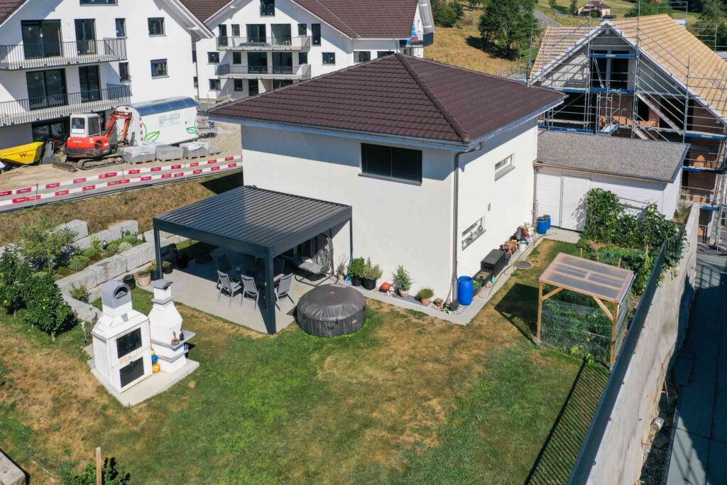 Luftaufnahme eines Vorstadtgartens mit überdachter Terrasse, Grillplatz und angrenzenden Häusern. Im Hintergrund sind Bauarbeiten und Fahrzeuge zu sehen.