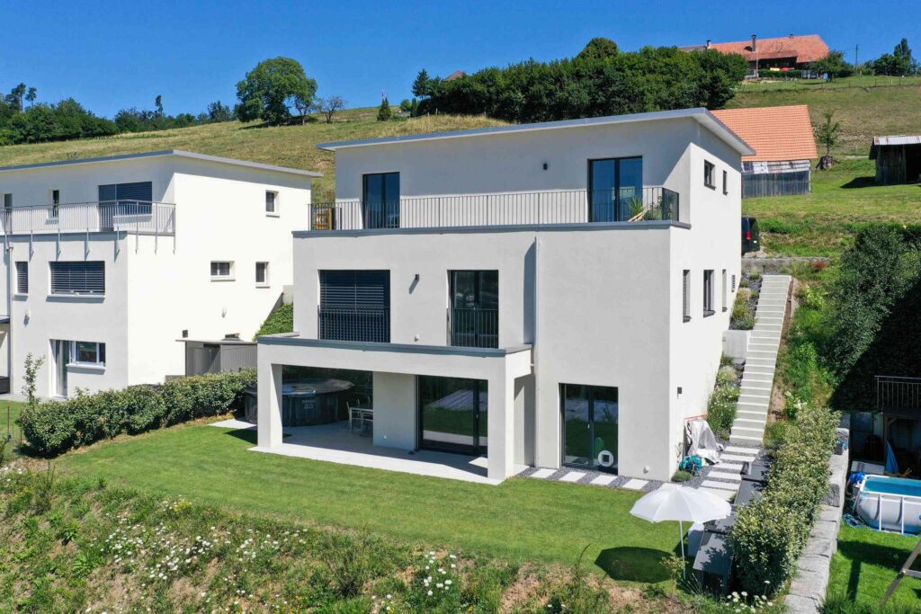 Modernes weißes zweistöckiges Haus mit Flachdach, großen Fenstern und Balkon. Umgeben von Rasen und Garten, mit Hügellandschaft im Hintergrund und einem Nachbarhaus auf der linken Seite.