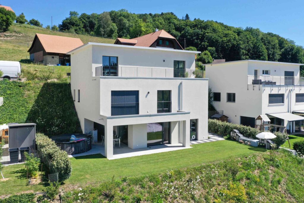 Ein modernes zweistöckiges Haus mit weißen Wänden und großen Fenstern auf einem grasbewachsenen Hügel, flankiert von ähnlichen Gebäuden und einer grünen Kulisse.