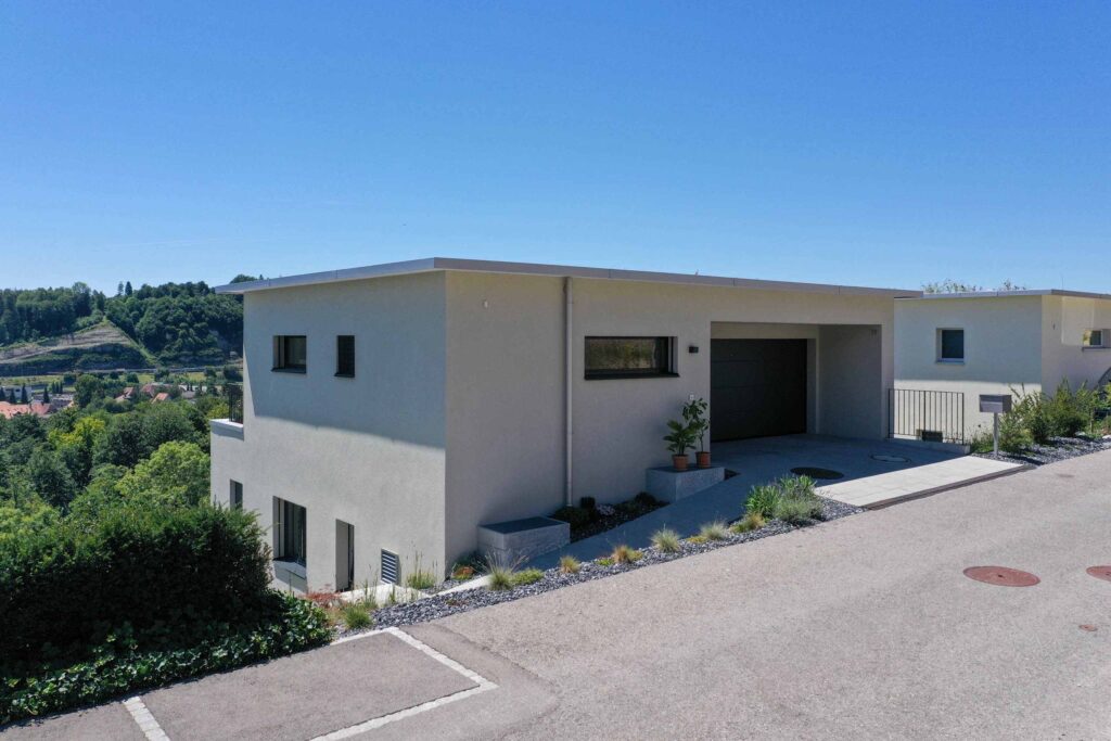Modernes weißes Haus mit Flachdach und Garage, an einem leichten Hang gelegen. Umgeben von Vegetation und einem klaren blauen Himmel.