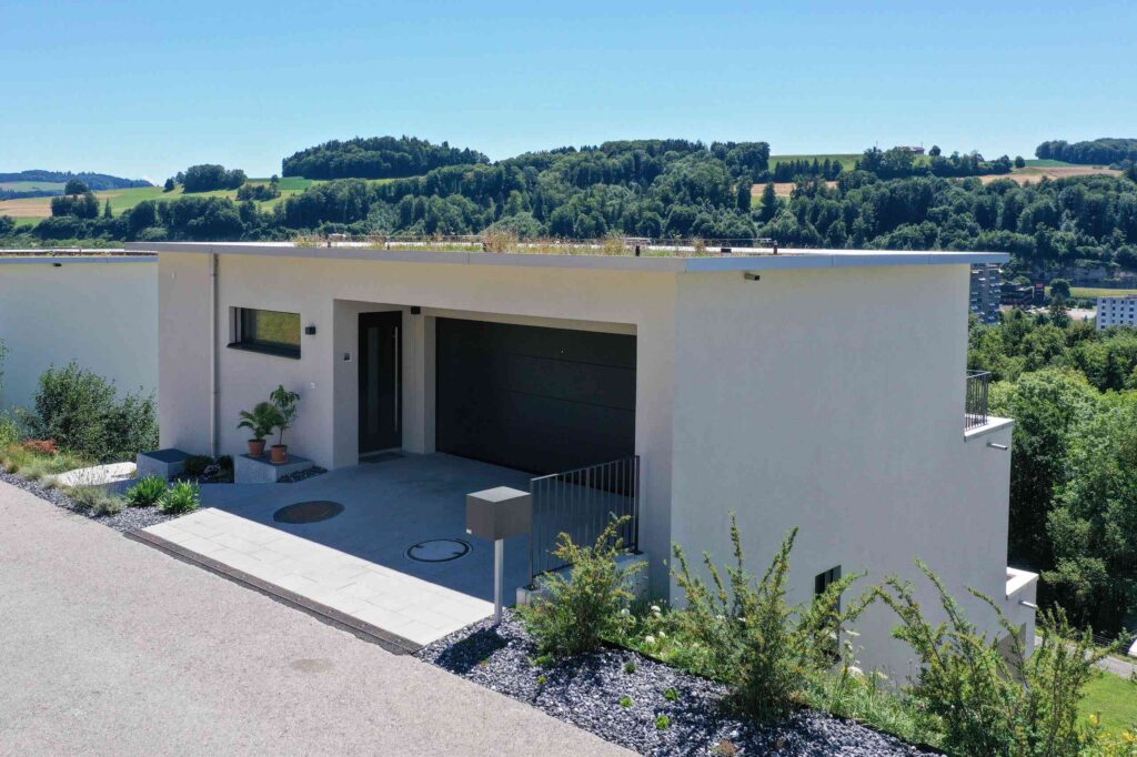 Modernes, weißes Haus mit Flachdach und Garage, an einem Hang gelegen, mit einer Naturlandschaft im Hintergrund unter einem klaren blauen Himmel.