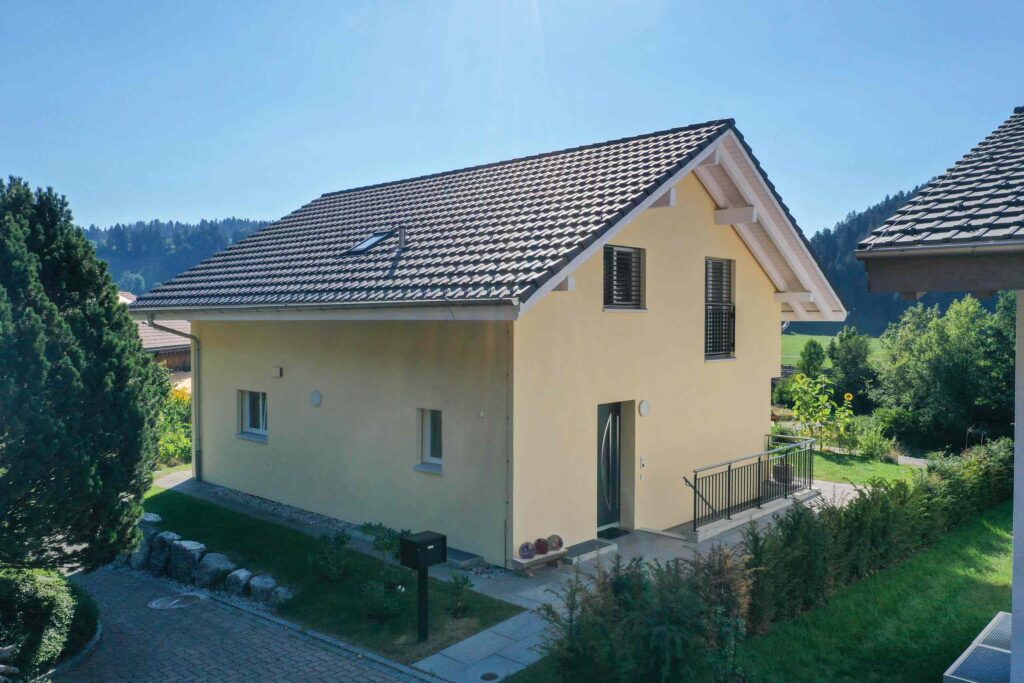 Kleines gelbes Haus mit schrägem Dach und zwei Fenstern, umgeben von Grün und Bäumen unter einem klaren blauen Himmel.