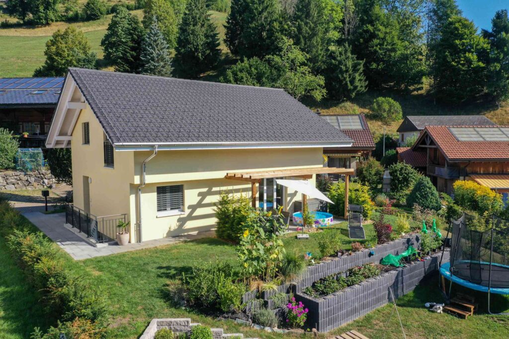 Ein zweistöckiges Haus mit Garten, Terrasse, Trampolin und Sonnenkollektoren. Umgeben von Bäumen und anderen Häusern in ländlicher Umgebung.