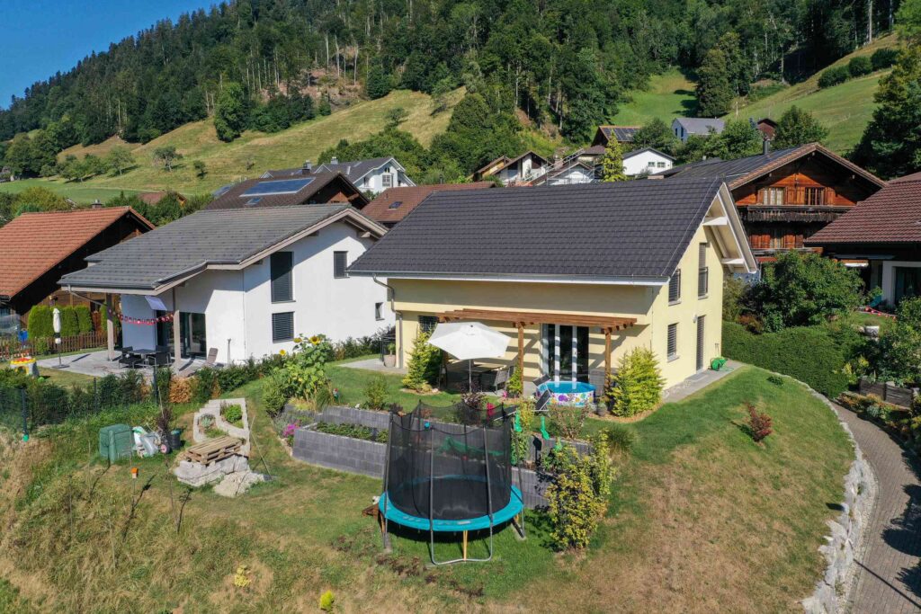 Luftaufnahme eines Hauses mit Garten, Trampolin und Sonnenschirm. Im Hintergrund sind andere Häuser und üppiges Grün zu sehen.