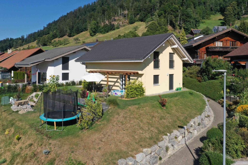 Ein Haus mit grauem Dach in Hanglage, umgeben von anderen Häusern, verfügt über ein Trampolin im Garten und überblickt eine abschüssige Rasenfläche mit einem steingesäumten Weg.