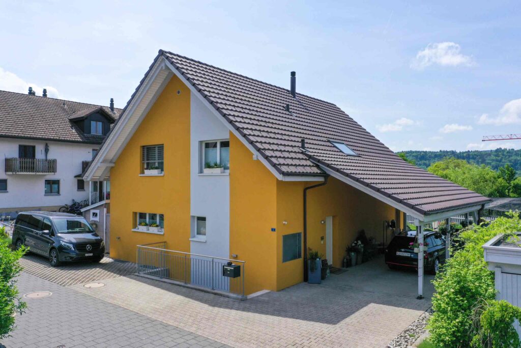 Ein gelbes Haus mit Schrägdach und einem Carport auf der rechten Seite, in dem ein Auto geparkt ist. Auf der Einfahrt parkt ein schwarzer Lieferwagen. In der Nähe befinden sich weitere Wohngebäude und Grünflächen.