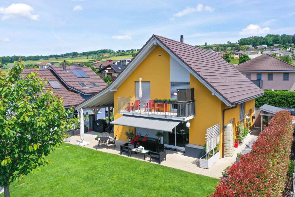 Ein gelbes Haus mit Schrägdach, Solarmodulen und Balkon. Der Hof ist mit grünem Gras und Hecken bewachsen. Im Hintergrund sind weitere Häuser unter blauem Himmel zu sehen.
