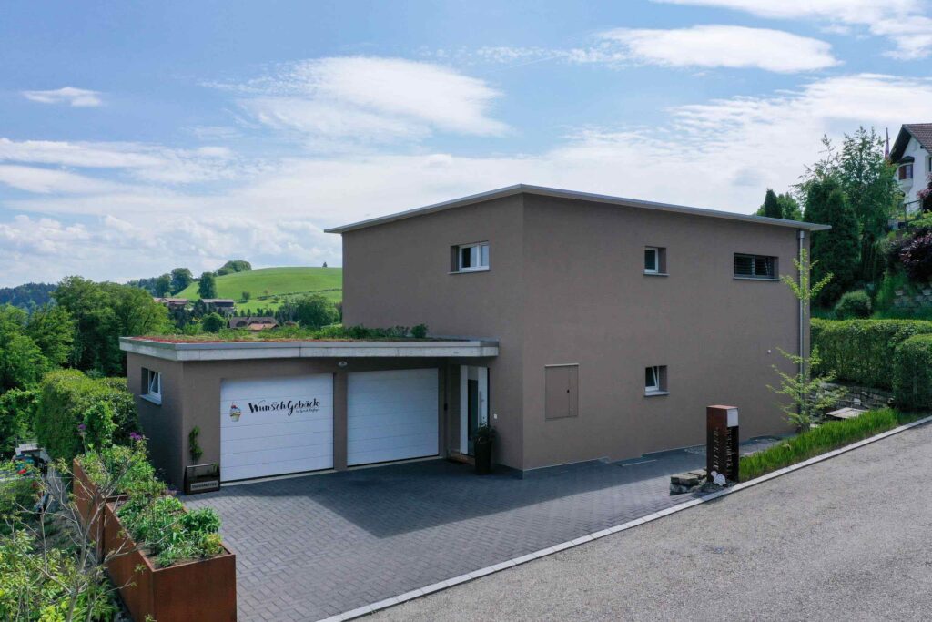 Ein modernes, zweistöckiges beiges Haus mit Flachdach, auf einem Hügel gelegen, umgeben von Grün, mit einer Auffahrt und einer Garage mit dem Logo „Vinici Architects“.